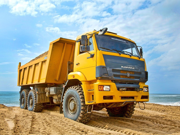 Жёлтый самосвал марки KAMAZ на песчаном карьере с голубым небом на фоне и видимым горизонтом моря.
